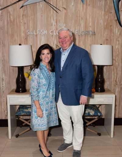 A woman and a man stand smiling in front of a wooden wall with "Sailfish Club 1914 Florida" written on it, beneath a mounted sailfish, as they attend the Annual Auction & Ball, framed by two lamps on a console table.