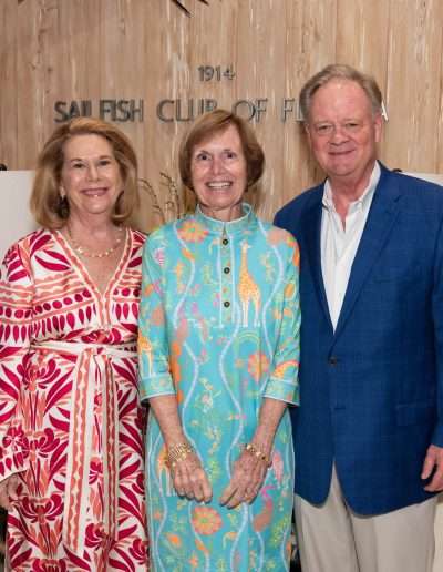 Three adults stand side by side smiling in front of a wooden wall with "Sailfish Club of Florida" signage, dressed elegantly for the Annual Auction & Ball; two women wear patterned dresses and a man sports a blue blazer.