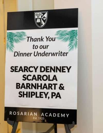 A sign reads “Thank You to our Dinner Underwriter Searcy Denney Scarola Barnhart & Shipley, PA” with the Rosarian Academy logo and name at the bottom for the Annual Auction & Ball.