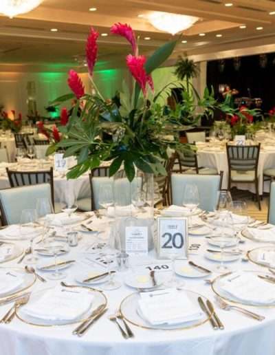 A banquet hall with round tables set for the Annual Auction & Ball, featuring white tablecloths, place settings, menus, and tall floral centerpieces with red flowers.