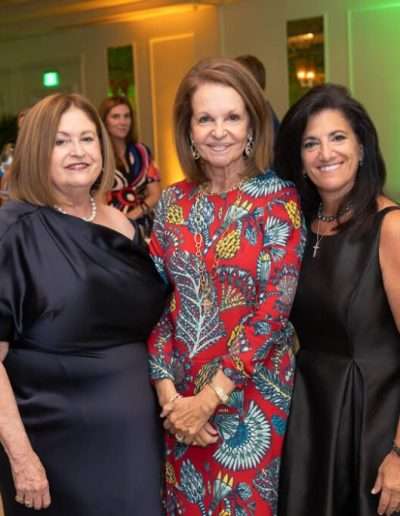 Three women pose together at the Annual Auction & Ball, dressed in formal attire. The background features other guests, green lighting, and elegant decor.