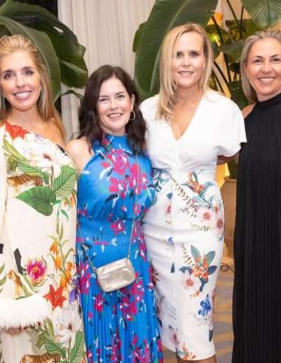 Four women stand indoors in front of large potted plants, wearing colorful, patterned dresses and smiling at the camera during the Annual Auction & Ball.