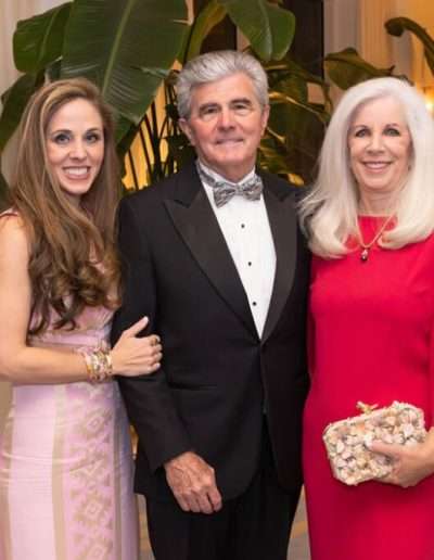 Three adults pose together indoors at the Annual Auction & Ball; a woman in a pink dress, a man in a black tuxedo, and a woman in a red dress holding a clutch. Large plants and lounge furniture are in the background.