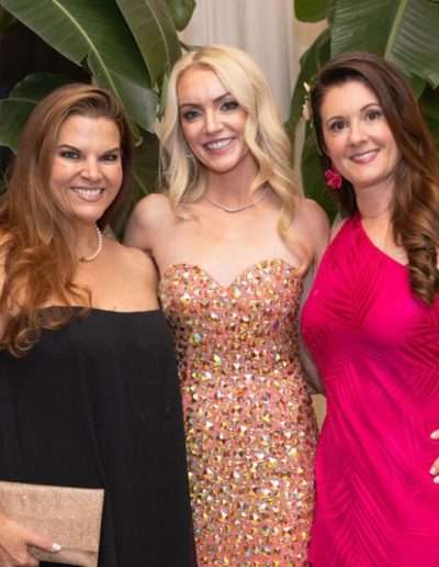Three women pose together indoors at the Annual Auction & Ball, dressed in formal evening wear, with large potted plants and soft lighting in the background.