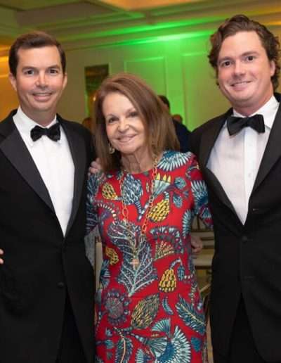 Three people pose for a photo at the Annual Auction & Ball; two men in black tuxedos stand on either side of a woman in a colorful patterned dress.
