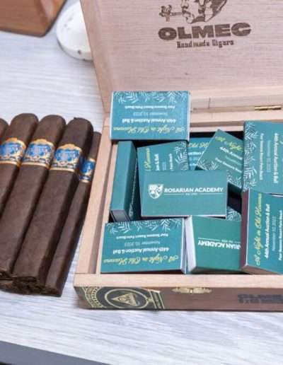 Five cigars are placed next to an open wooden box filled with green matchboxes labeled "ROSARIAN ACADEMY," set on a light-colored surface—perfect for celebrating the Annual Auction & Ball.