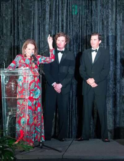 At the Annual Auction & Ball, four people in formal attire stand on stage; a woman at a podium speaks as two men in tuxedos stand beside her and another woman in a red dress applauds.
