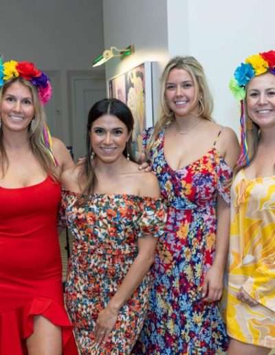 Four women in colorful dresses and floral headpieces stand together indoors, smiling at the camera during the Annual Auction & Ball.